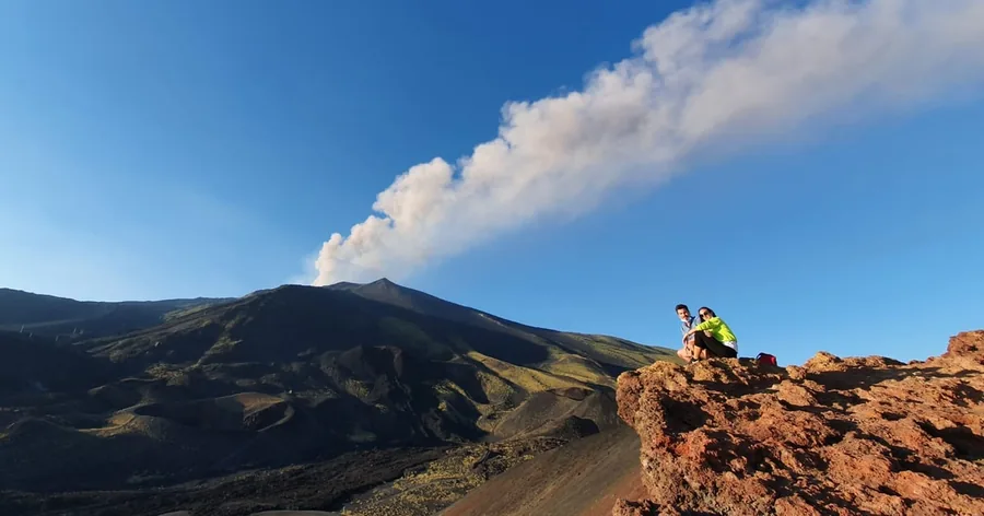 Un pennacchio di gas bianco che si alza dalla cima dell'Etna sopra versanti scuri, un escursionista su uno sperone di roccia rossa