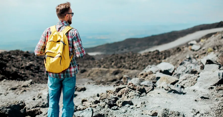 Un escursionista con lo zaino giallo che guarda le distese di lava grigia dell'Etna verso il mare
